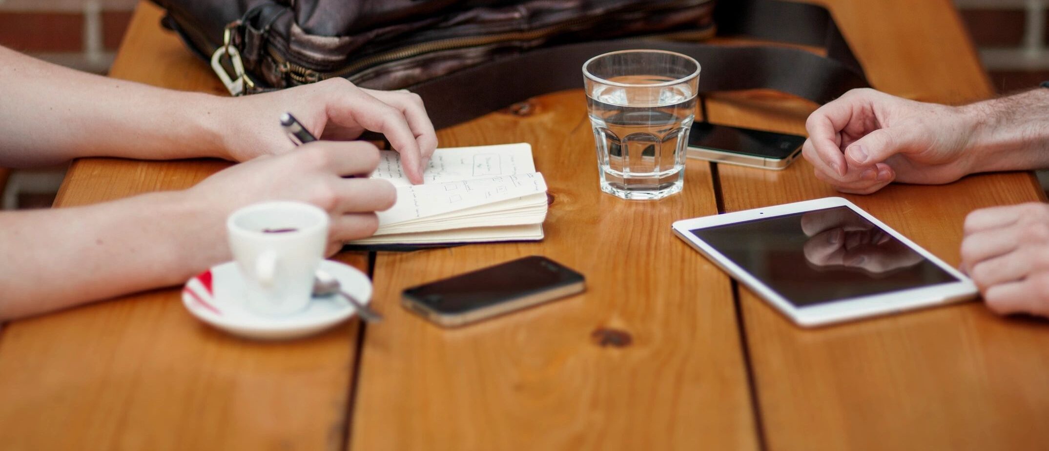 background imagery showing the hands of two people sitting opposite each other on a table. There's a bag along with a coffee mug, a glass or water, two phones, and a tablet. One person is jotting notes in a notebook.